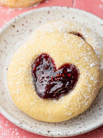 Thumbprint cookie with a heart shape indent on a white plate with powdered sugar.