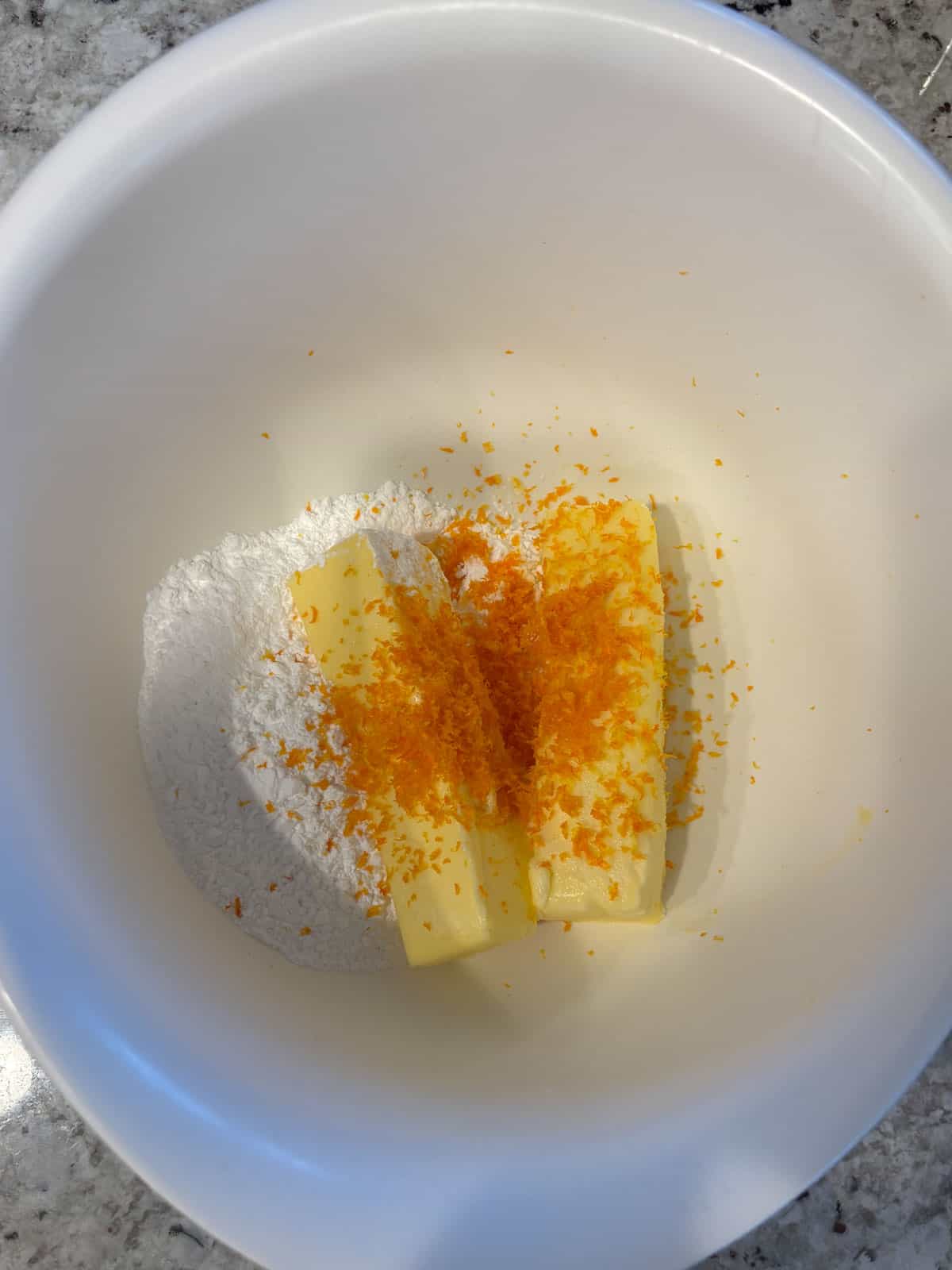 Powdered sugar, orange zest, and butter in a white bowl ready to be mixed.