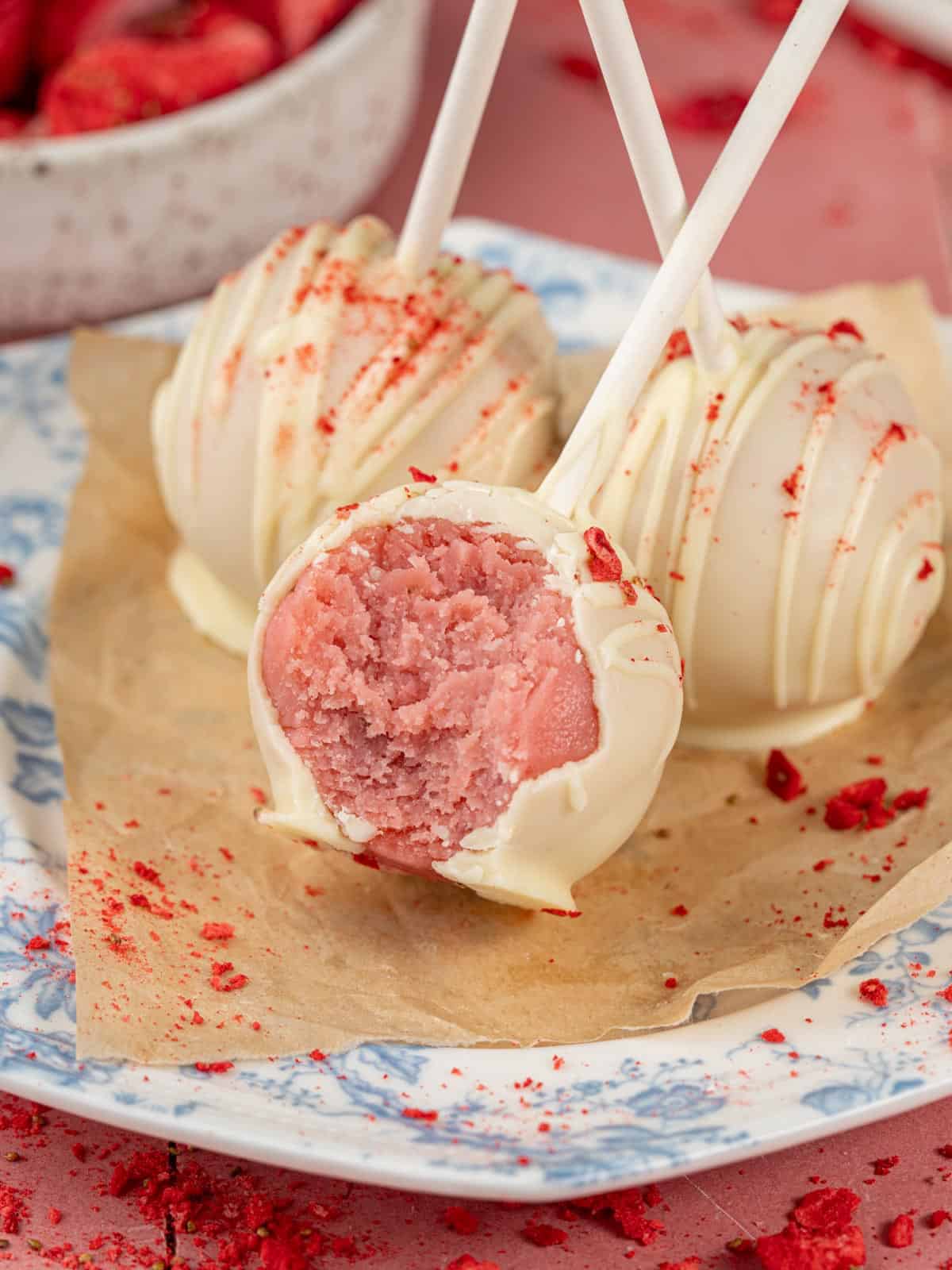 Strawberry cake pops with icing on a plate with strawberries around.