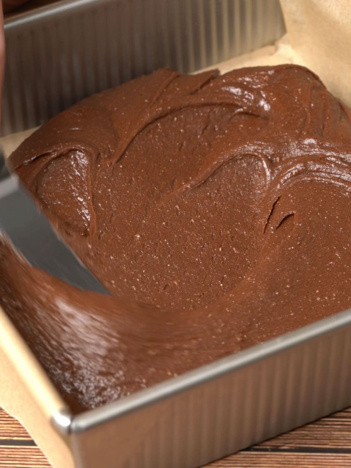 Brownie batter being spread into an 8X8 baking pan with parchment paper.
