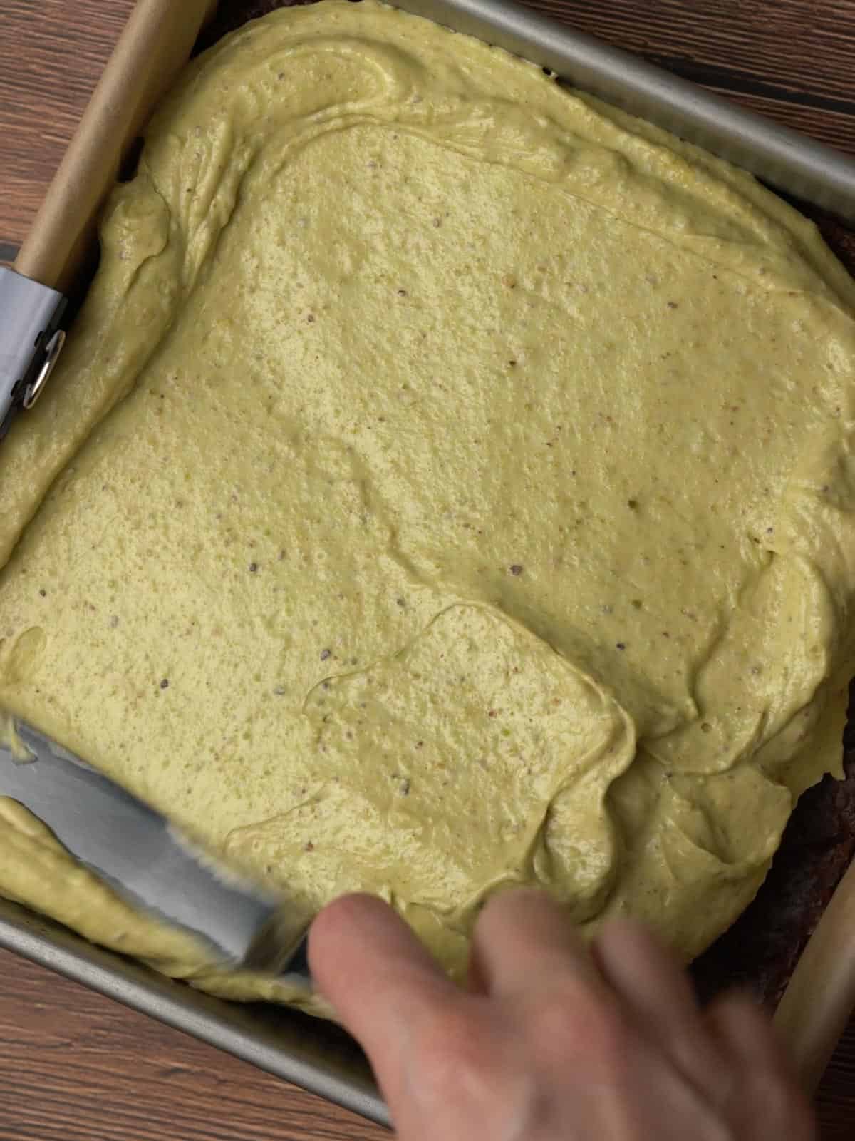 The pistachio layer being spread on top of the brownie in a pan.