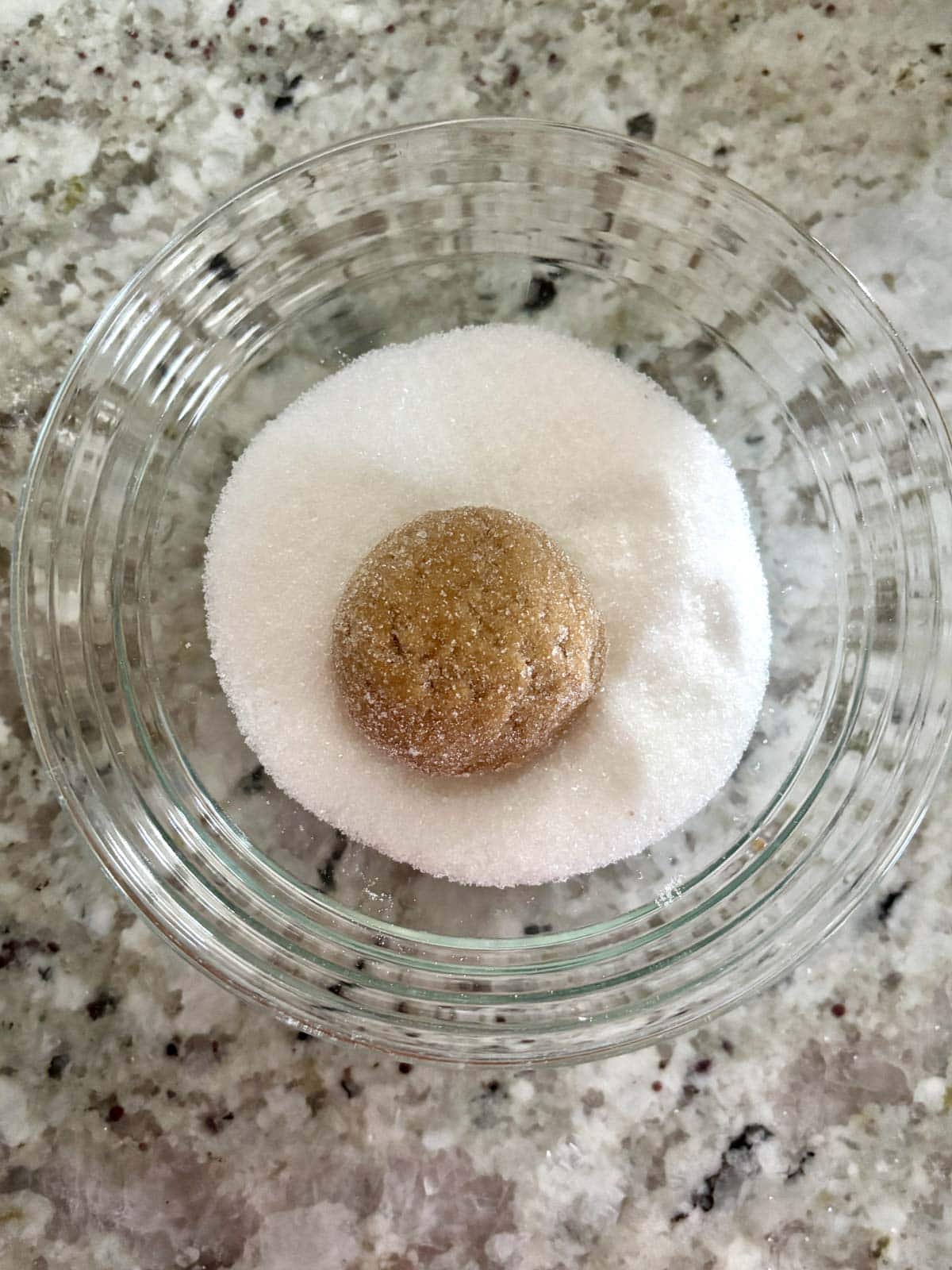 A ball of cookie dough being rolled in granulated sugar in a bowl.