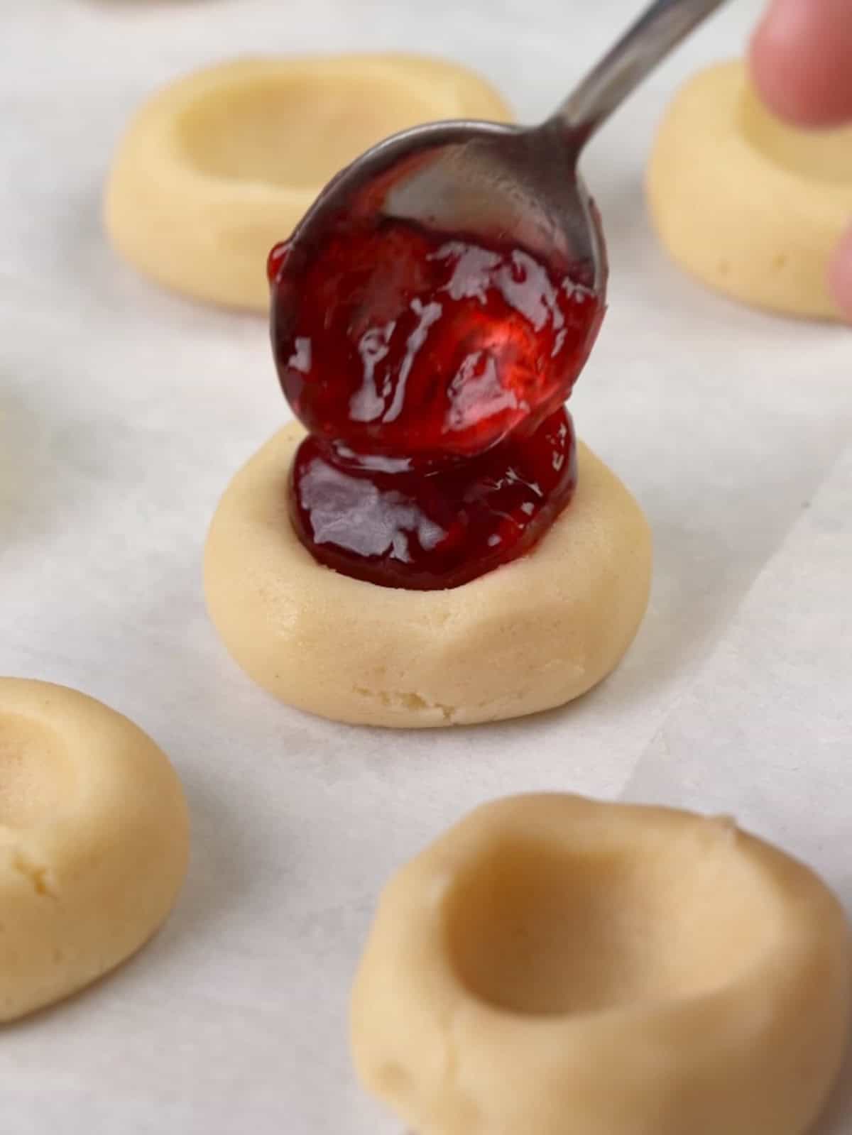 Raspberry jam being spooned into the cookie dough indent.