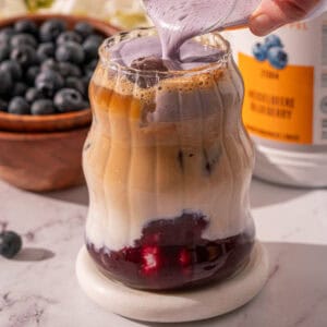 Pouring blueberry cold foam over the iced latte on a coaster.