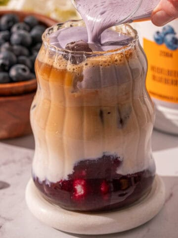 Pouring blueberry cold foam over the iced latte on a coaster.