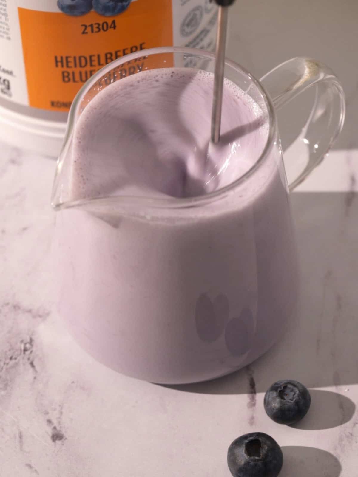 Frothing cream and blueberry syrup in a glass pitcher.