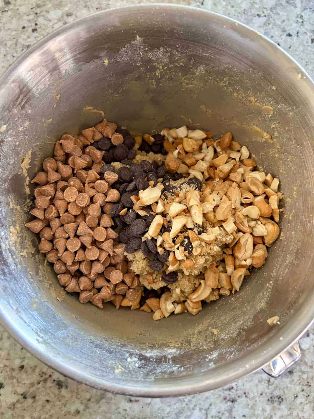 Butterscotch chips, chocolate chips, and cashews being added to the cookie dough in a mixing bowl.