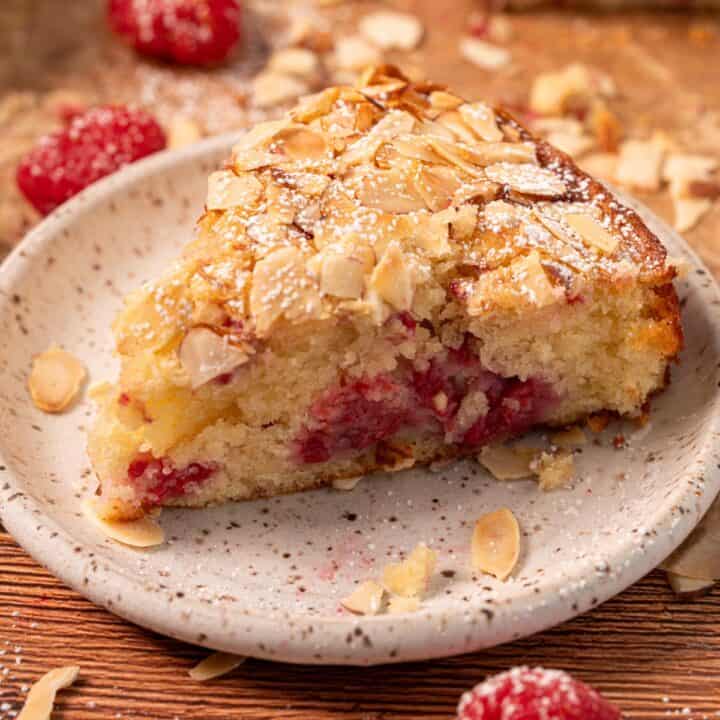 A slice of raspberry and almond cake on a plate with sliced almonds and fresh raspberries around.