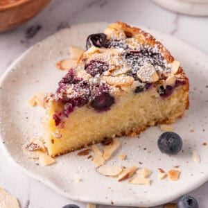 A slice of blueberry almond cake on a white plate with fresh blueberries and shaved almonds.