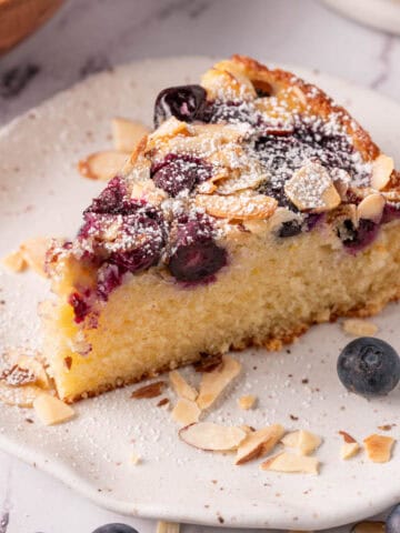 A slice of blueberry almond cake on a white plate with fresh blueberries and shaved almonds.