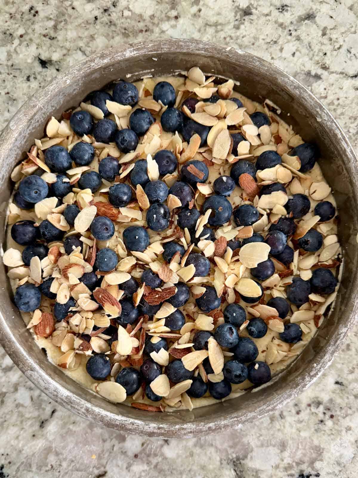 Blueberries and sliced shaved almonds on top of the almond cake batter in an 8 inch round cake pan.