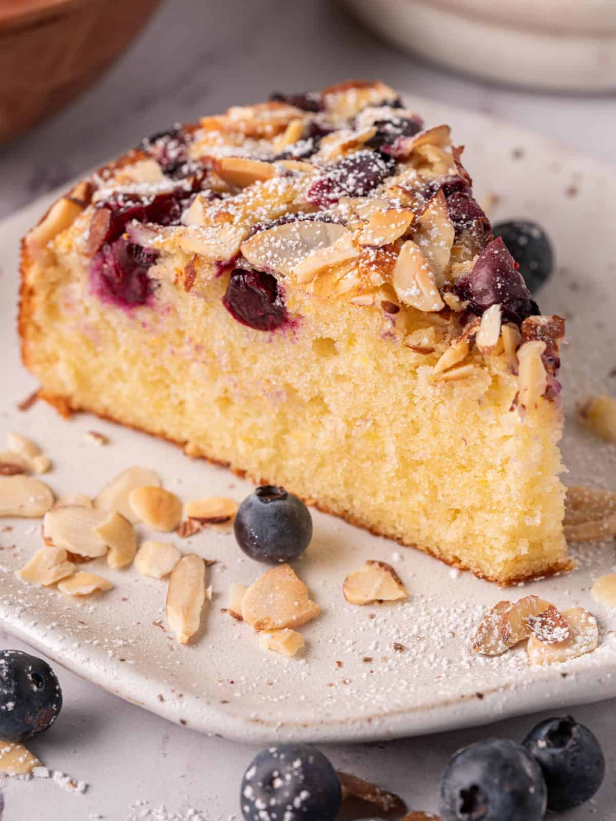 An old fashioned cake slice on a white plate showing the crumb, fresh blueberries, and sliced almonds.