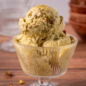 Pistachio nut ice cream in a glass dish with bowls in the background and pistachios on top.
