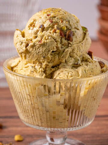 Pistachio nut ice cream in a glass dish with bowls in the background and pistachios on top.