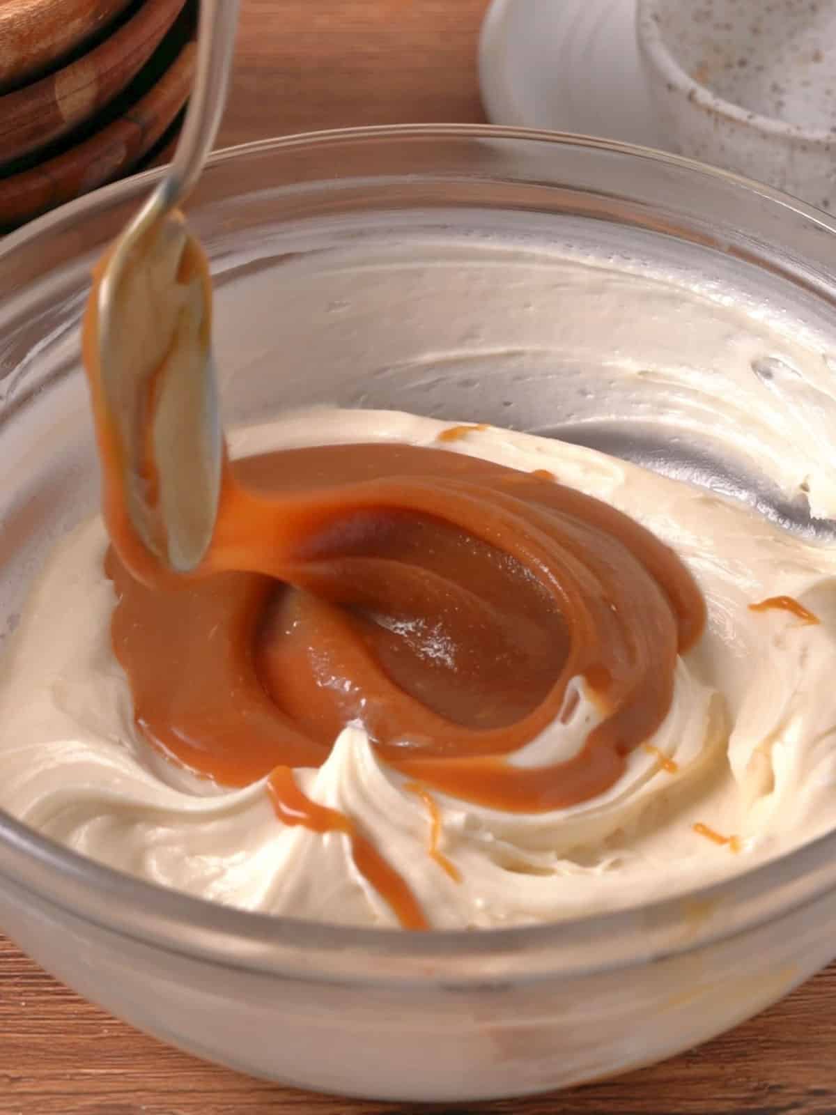 Adding salted caramel sauce to cream cheese frosting in a bowl on the counter.